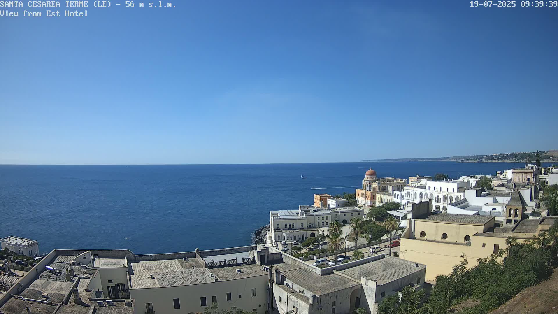 A coastal town with white buildings and a domed structure is situated next to a calm, blue sea under a clear, sunny sky.