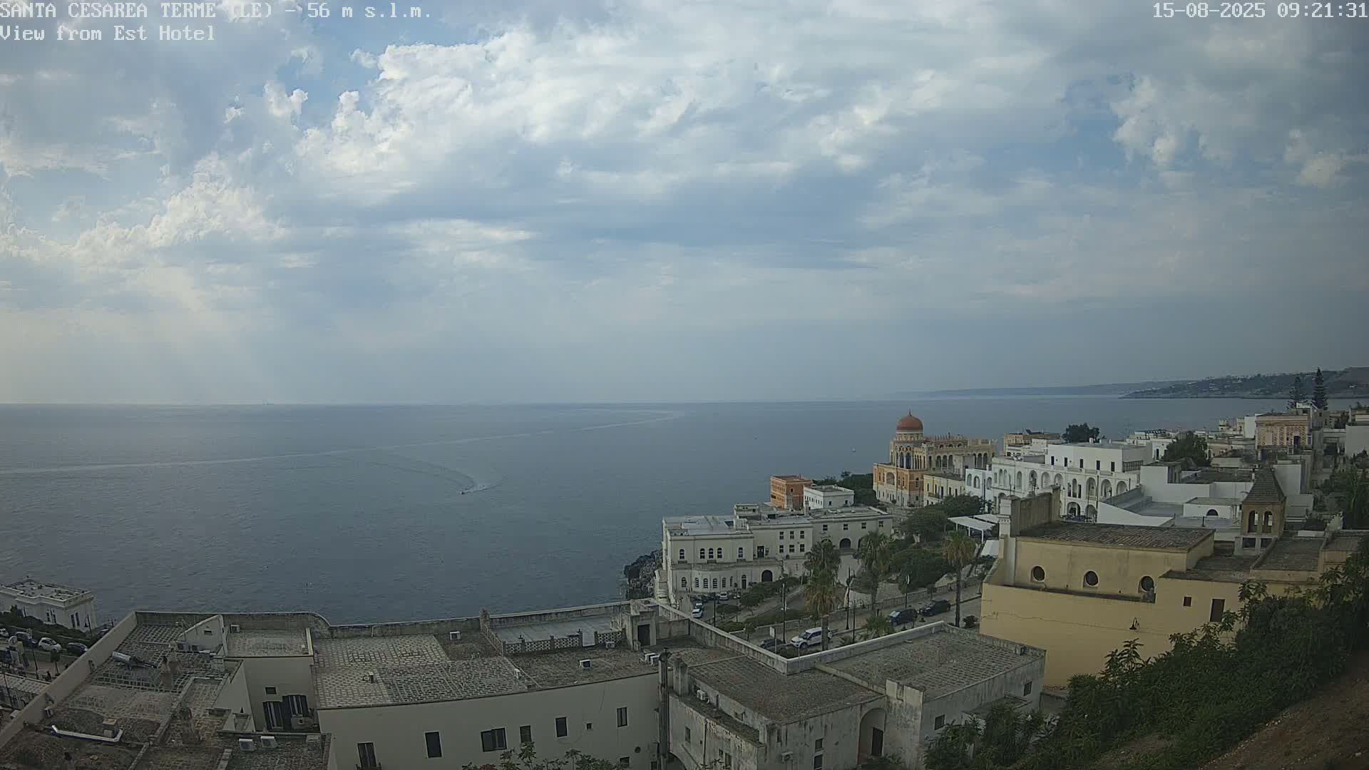 A coastal town with white buildings and a prominent domed structure overlooks a calm sea under a partly cloudy sky.