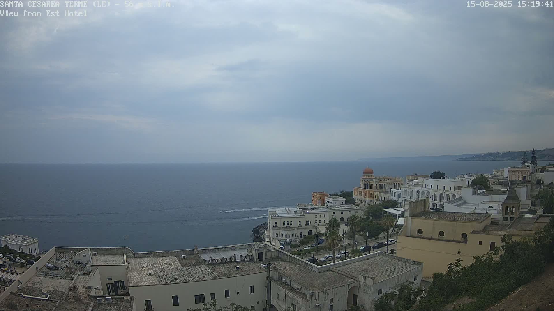 A coastal town with white buildings and a prominent domed structure overlooks a calm sea under a partly cloudy sky.