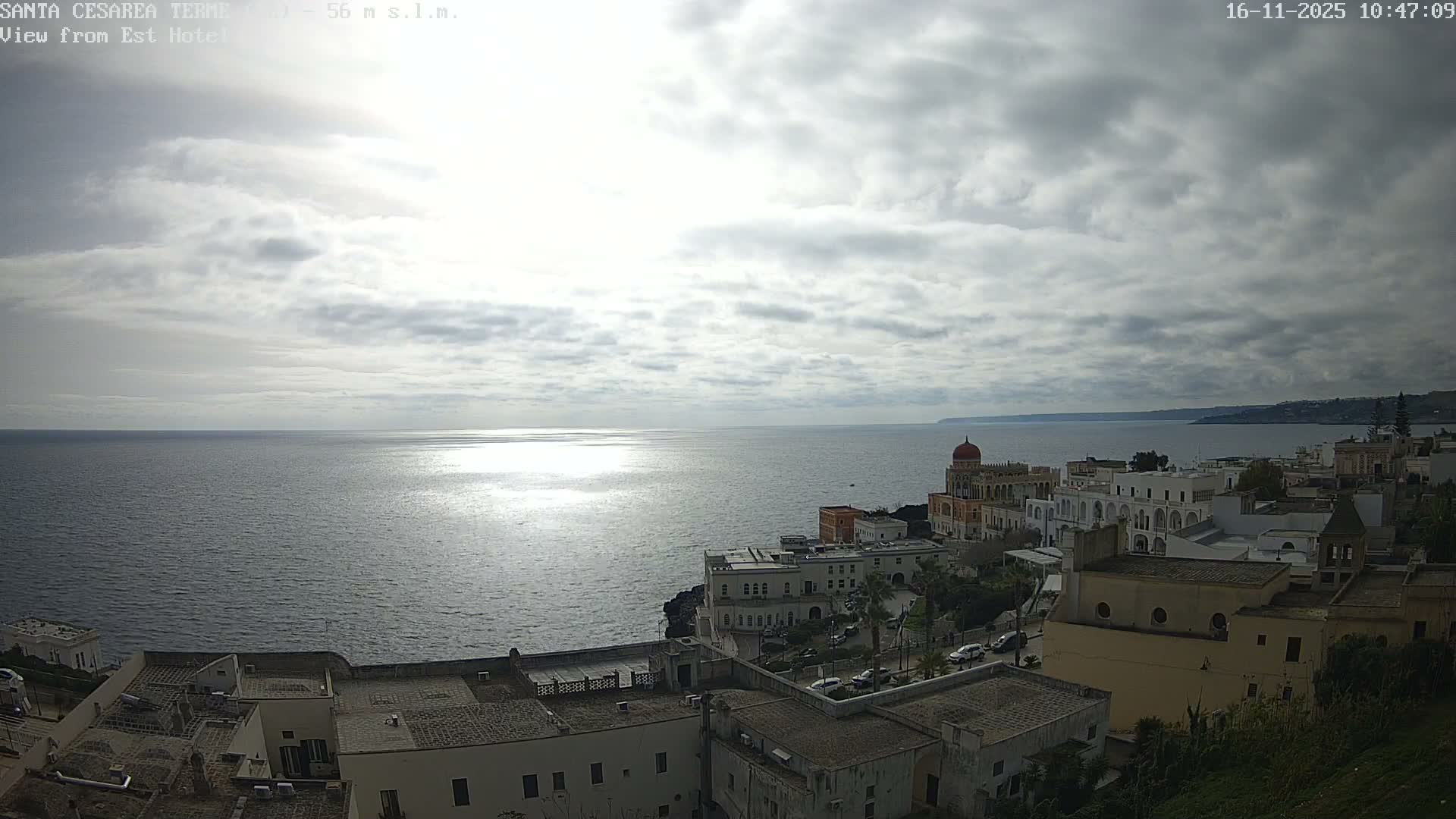 An overcast day overlooks a coastal town featuring a mix of white and historical architecture built into cliffs, with sunlight reflecting off the calm sea stretching to the horizon.