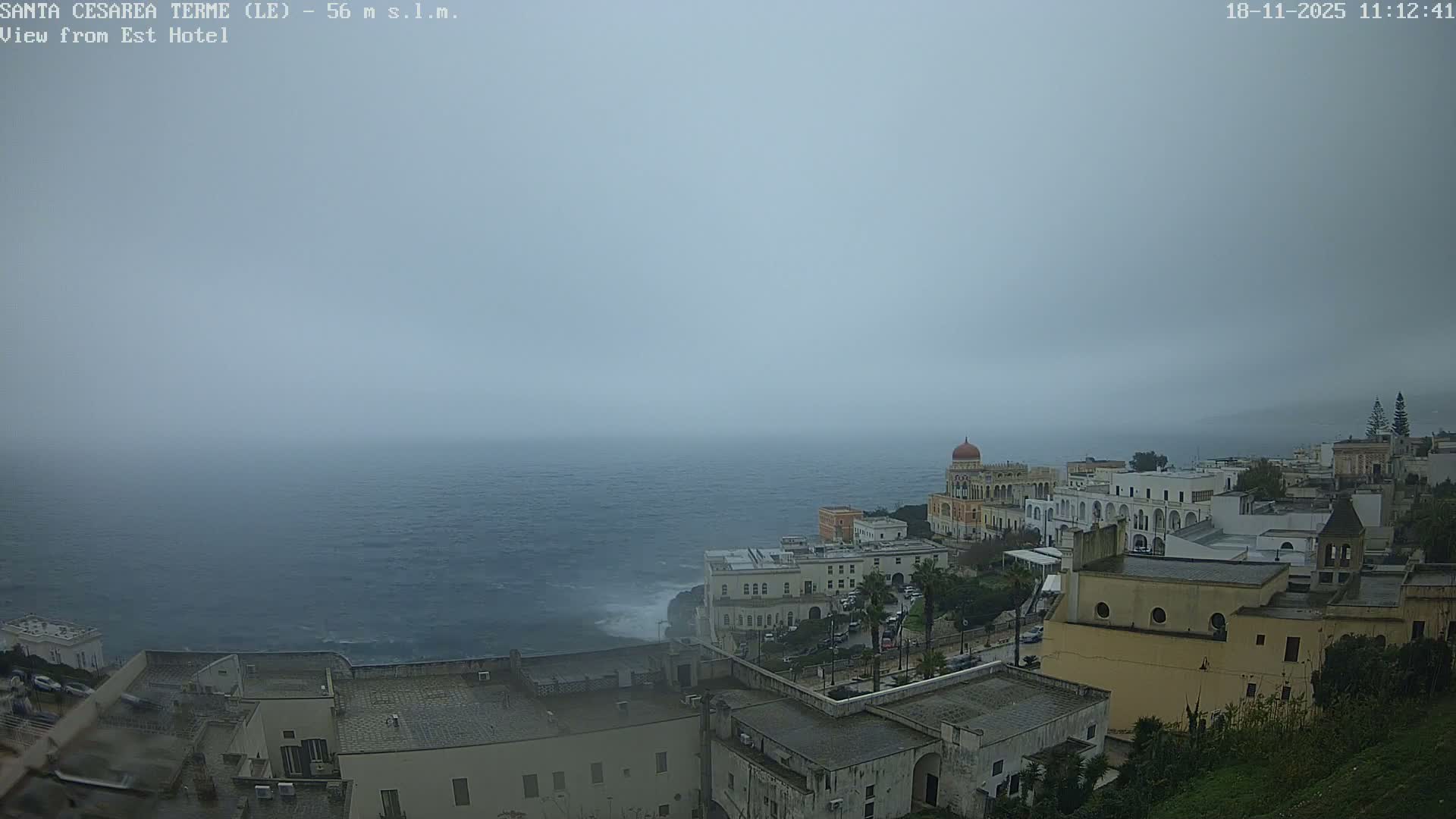 An overcast day overlooks a coastal town featuring a mix of white and historical architecture built into cliffs, with sunlight reflecting off the calm sea stretching to the horizon.