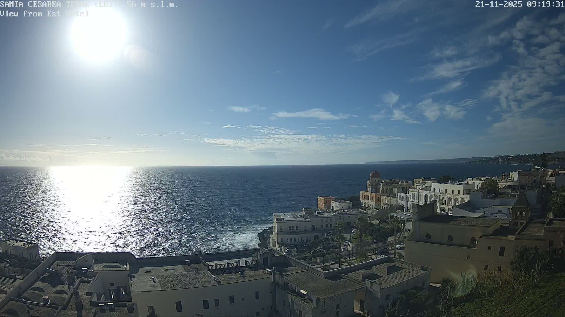 A bright, sunny day illuminates a Mediterranean coastal town, featuring white and ornate buildings perched on cliffs overlooking a sparkling blue sea under a sky with scattered clouds.