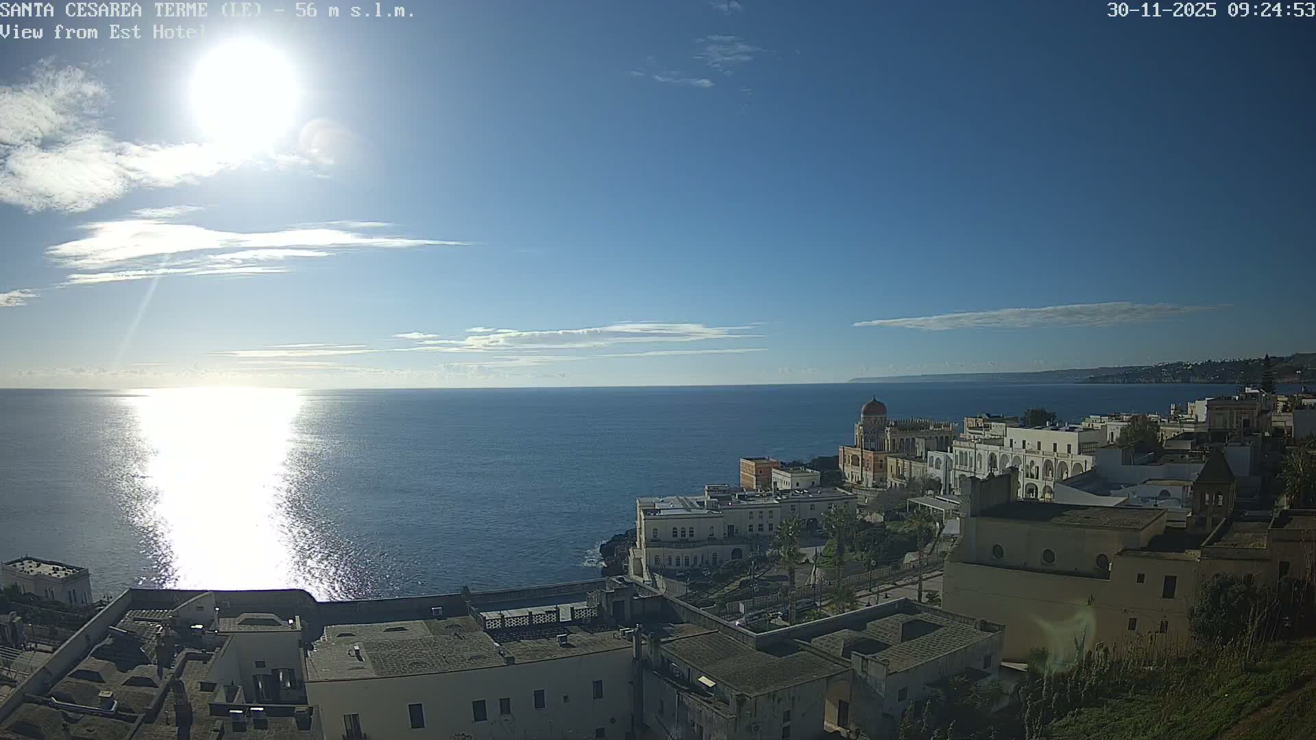 A panoramic view reveals a coastal town with buildings nestled along cliffs overlooking a calm, sun-drenched sea under a bright blue sky with scattered clouds.