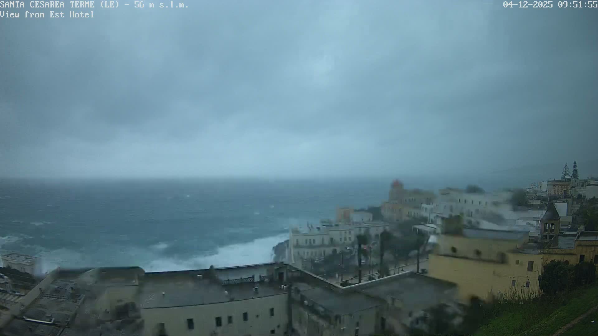 A stormy, overcast day shows strong waves crashing against the rocky coastline of a town with buildings lining the cliffs.
