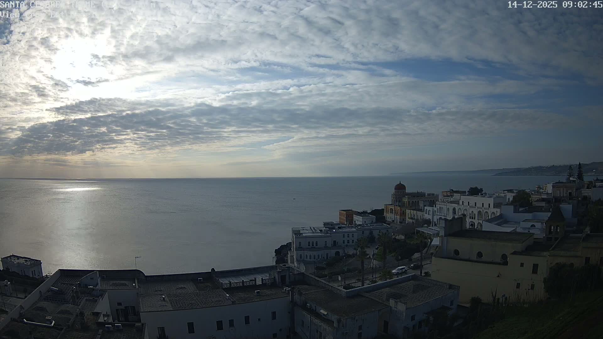 A coastal town with historic buildings overlooks a calm sea under a partly cloudy sky with bright sun reflecting on the water.