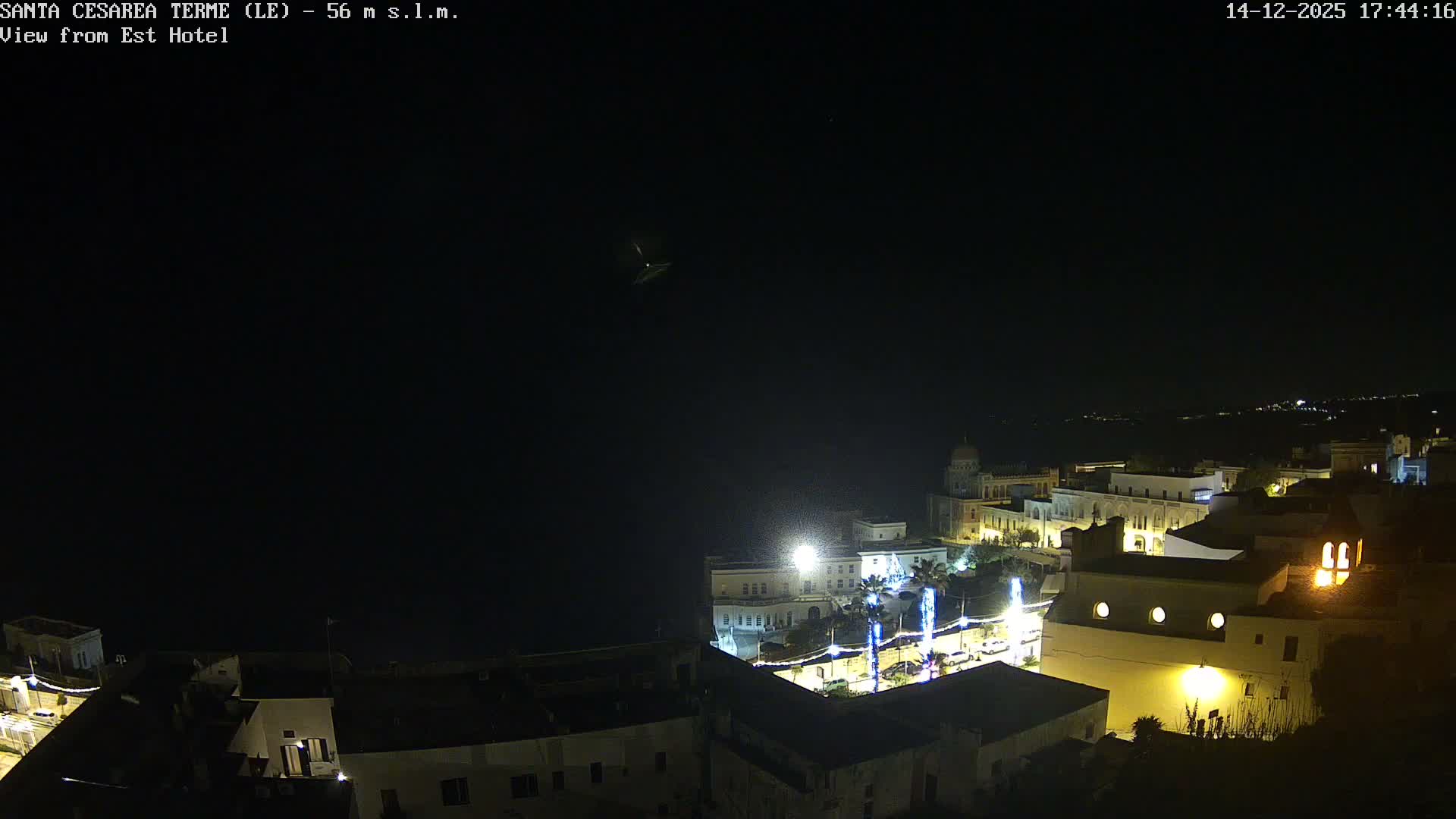 A coastal town with historic buildings overlooks a calm sea under a partly cloudy sky with bright sun reflecting on the water.