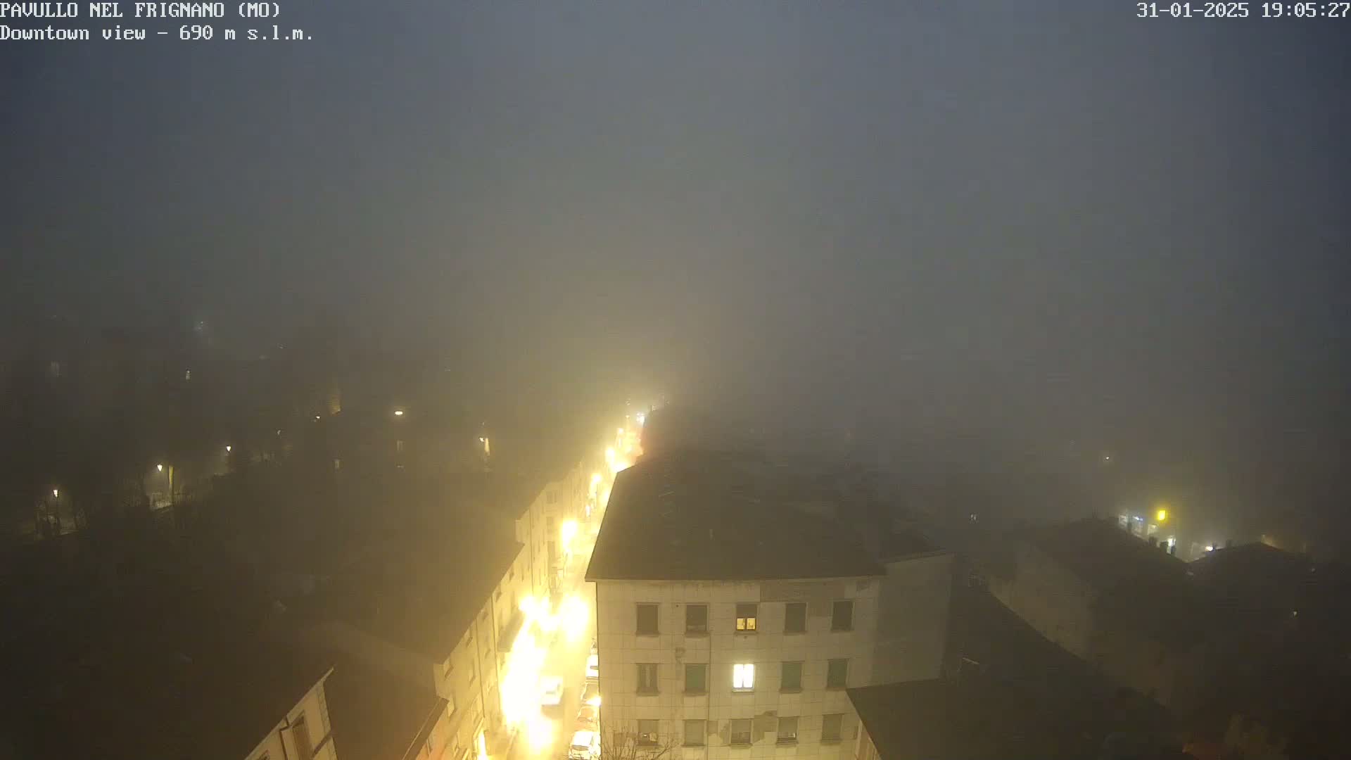 A foggy night scene shows a town's street lights illuminating buildings and a road below, seen from an elevated vantage point.