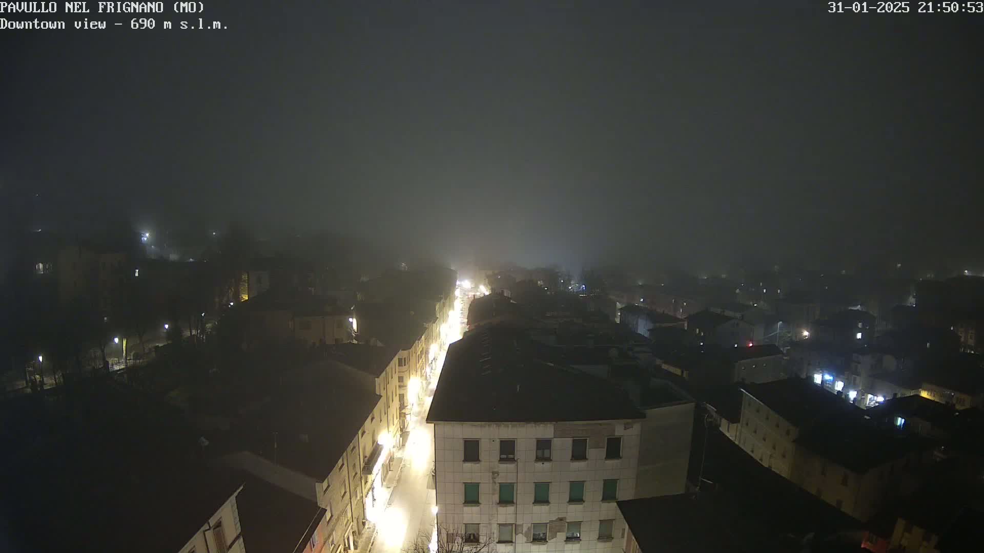 A foggy night scene shows a town's illuminated streets and buildings from an elevated viewpoint.