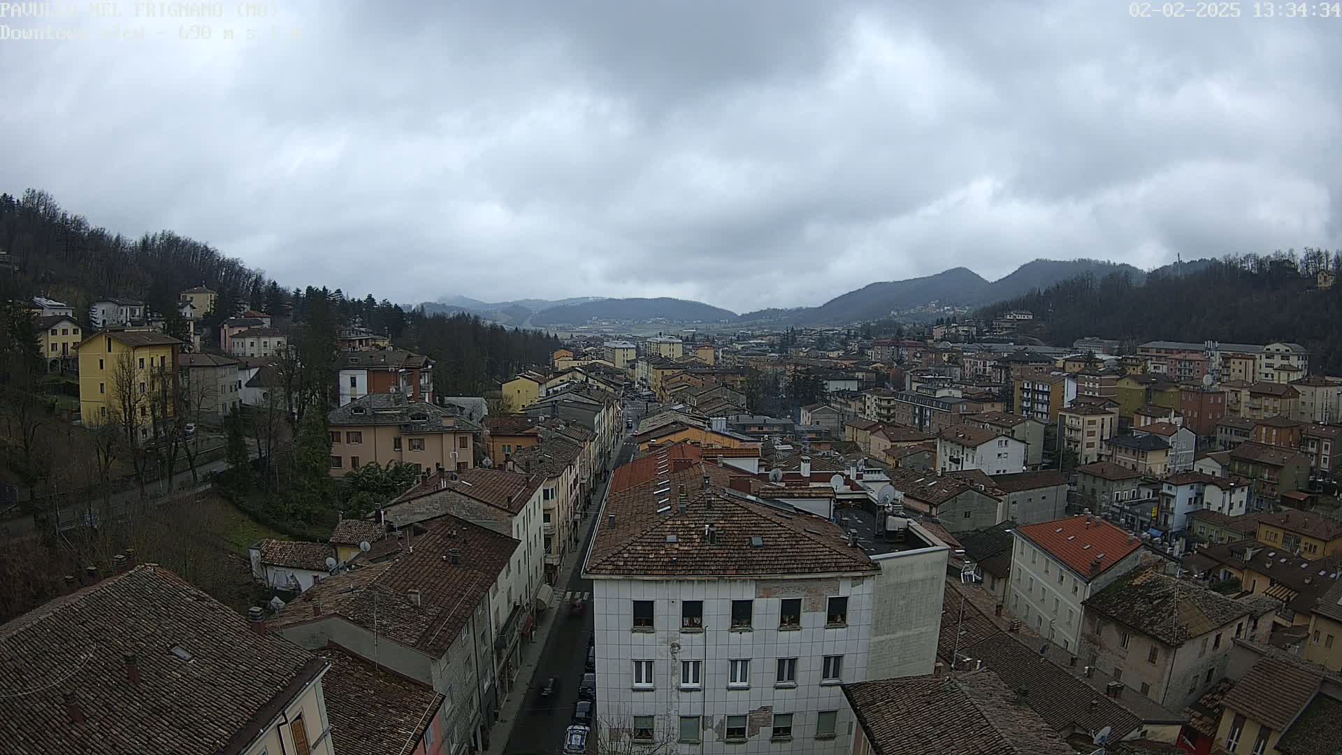 An elevated view of a town nestled in a valley under a cloudy sky, showing numerous buildings with red tile roofs and a street running through the center.