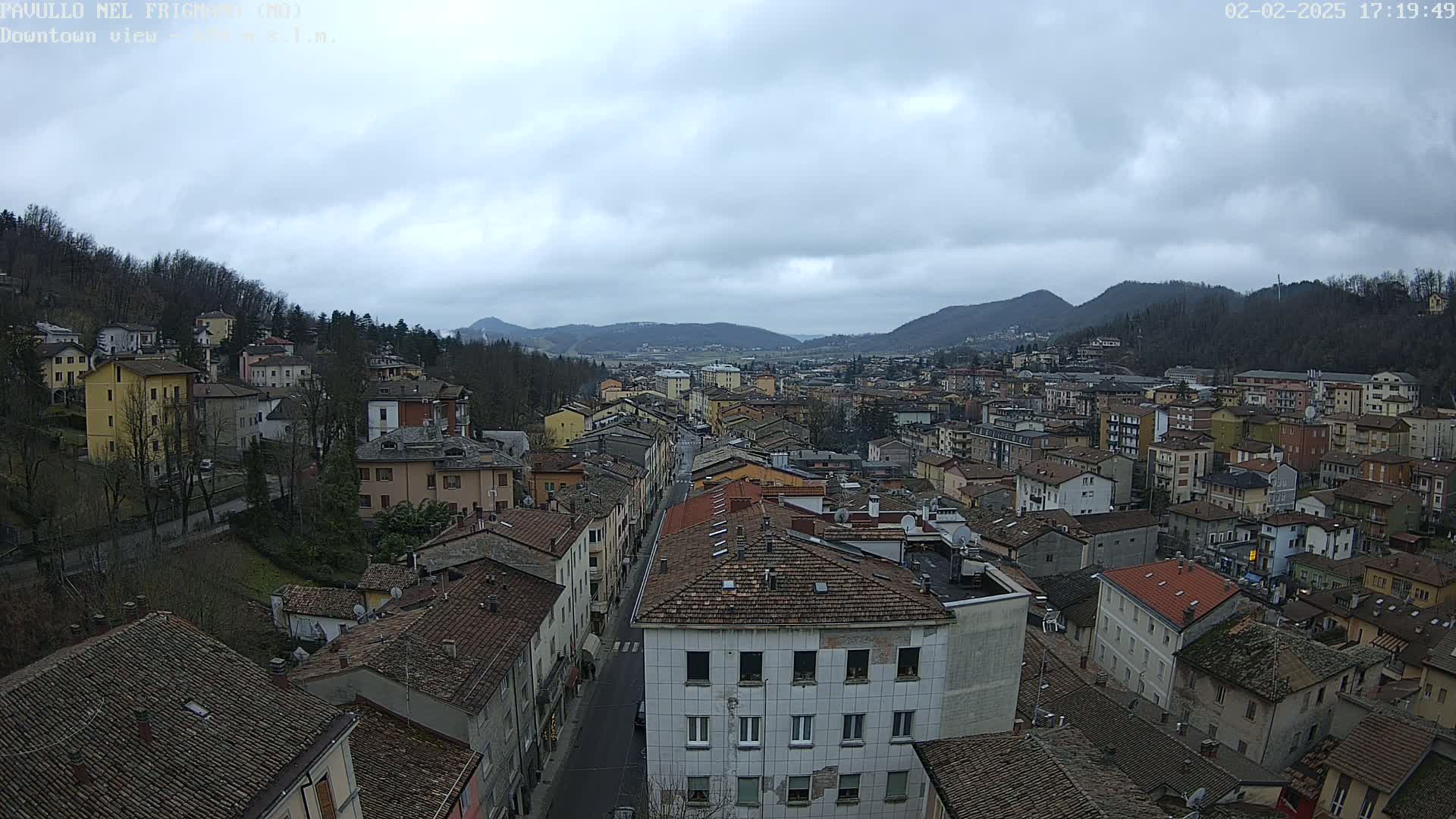 A view of a town nestled in a valley under an overcast sky, with many buildings with brown roofs and a street running through the center.