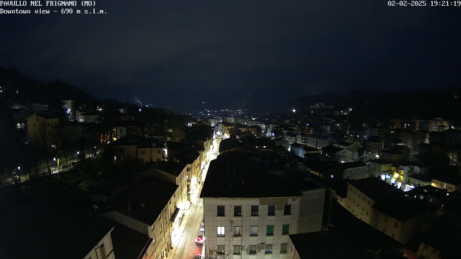 A nighttime, high-angle view of a town nestled in a valley, with a street illuminated by streetlights and a few car headlights visible.