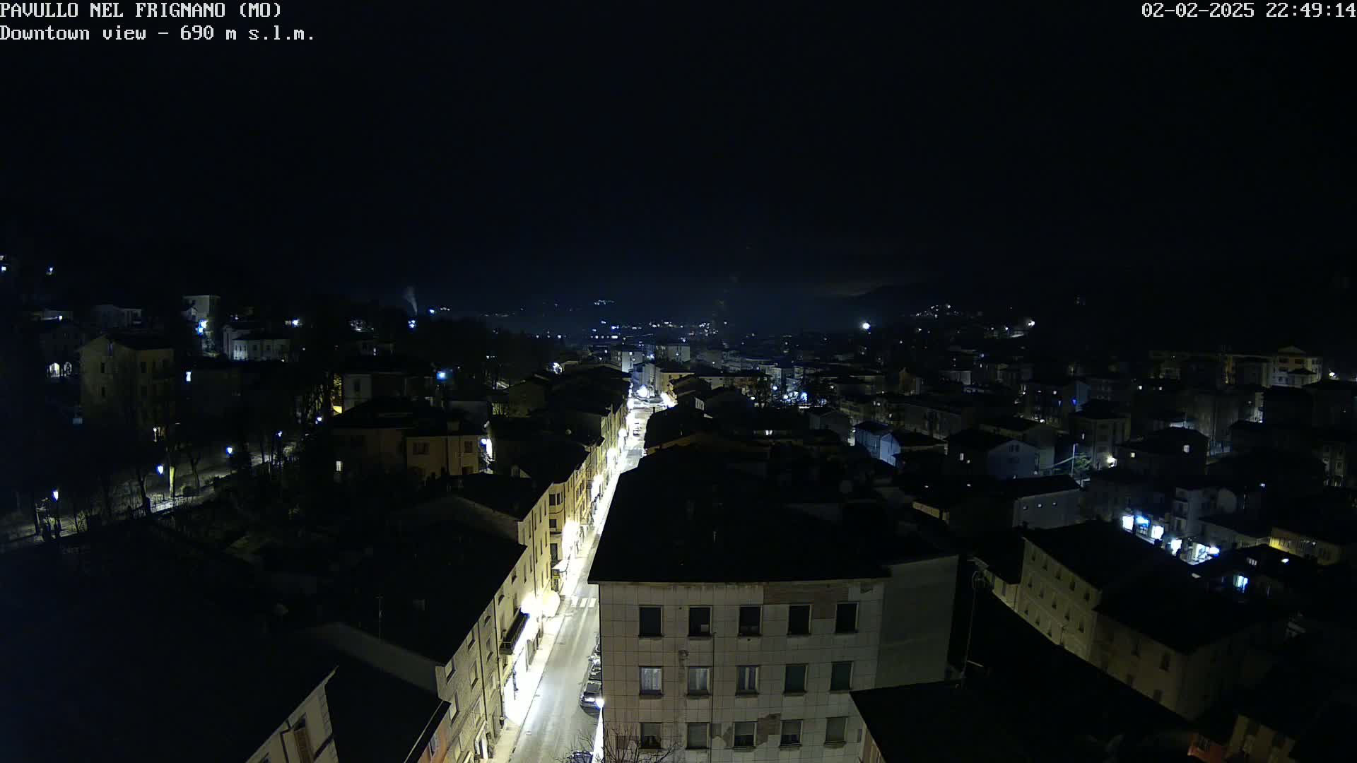 A nighttime aerial view of a town shows a long, illuminated street cutting through a dark expanse of buildings under a clear, starless sky.