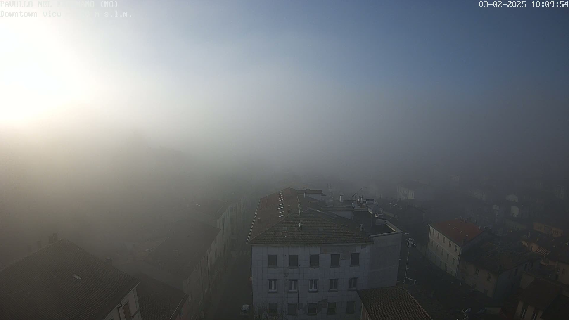 A thick fog obscures a city view, showcasing several buildings with rooftops visible above the dense mist under a mostly clear sky.