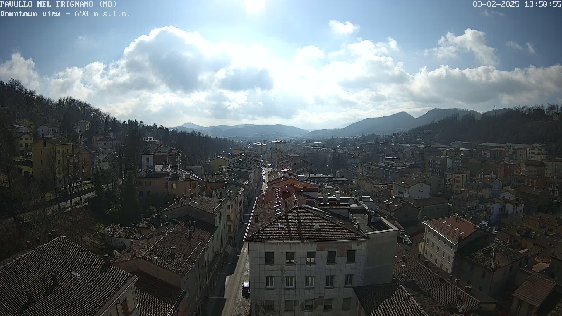 A partly sunny day reveals a sprawling town nestled in a valley between hills, with a long street lined with buildings visible from an elevated vantage point.