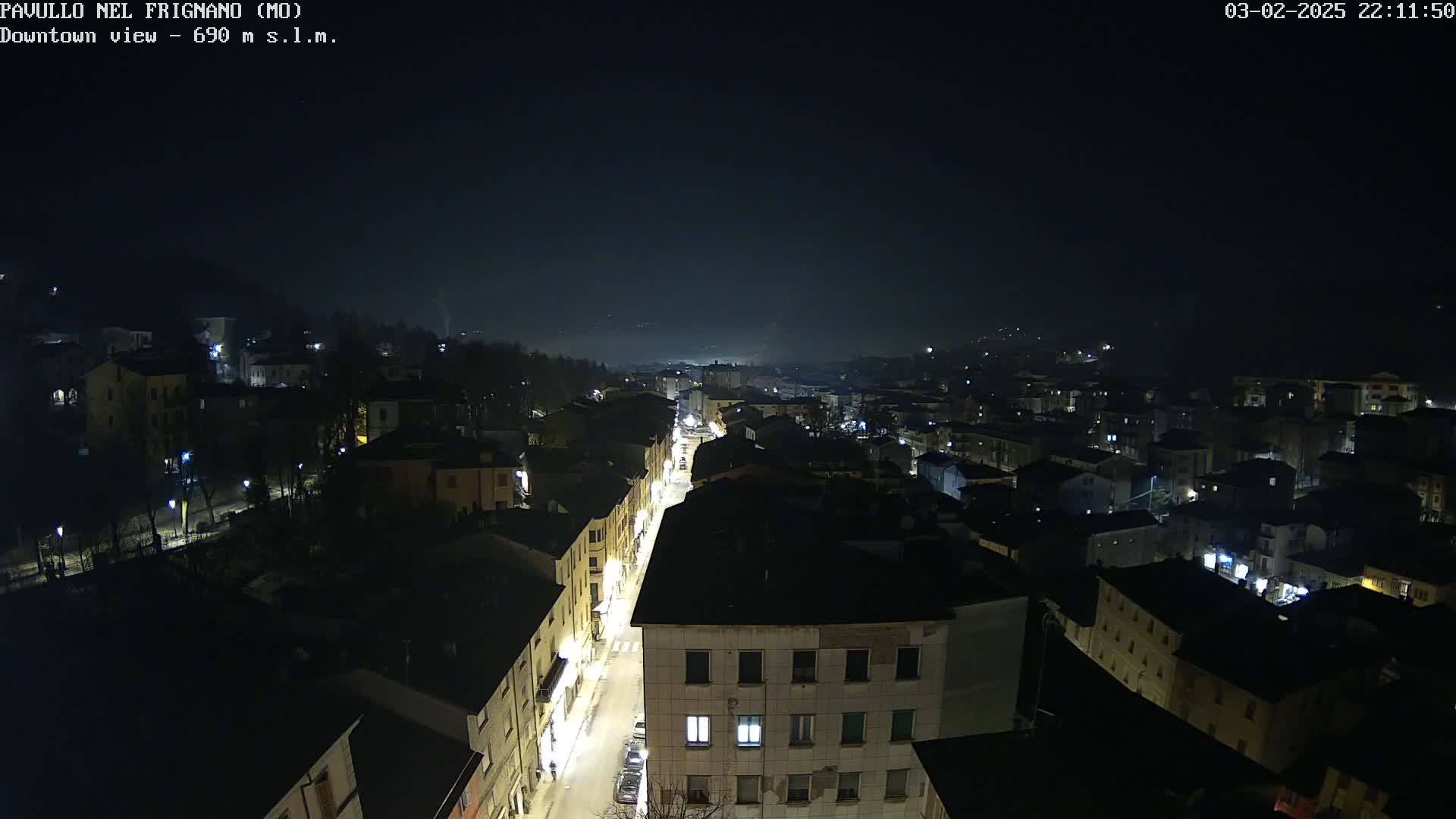 A nighttime aerial view of a town shows a long street with illuminated buildings on either side, under a dark, slightly hazy sky.