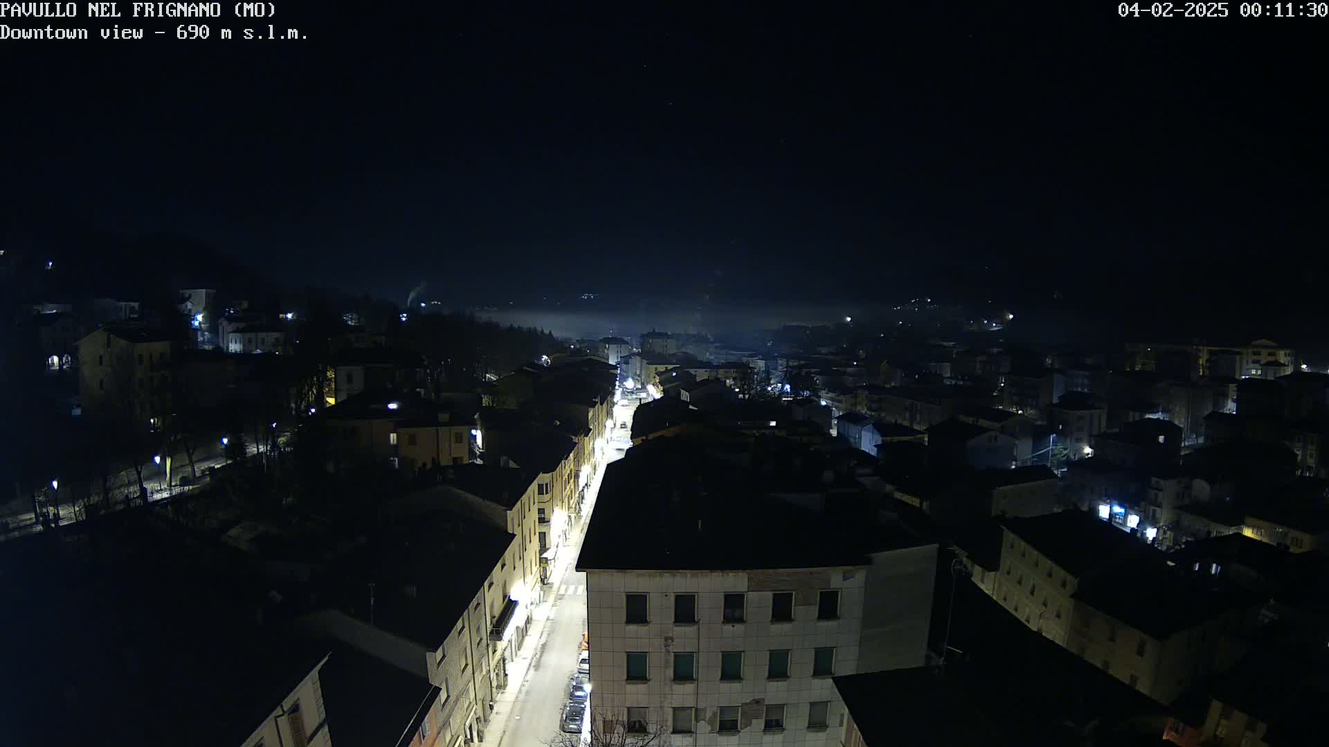 An aerial nighttime view of a town with a street illuminated, under a dark, slightly foggy sky.