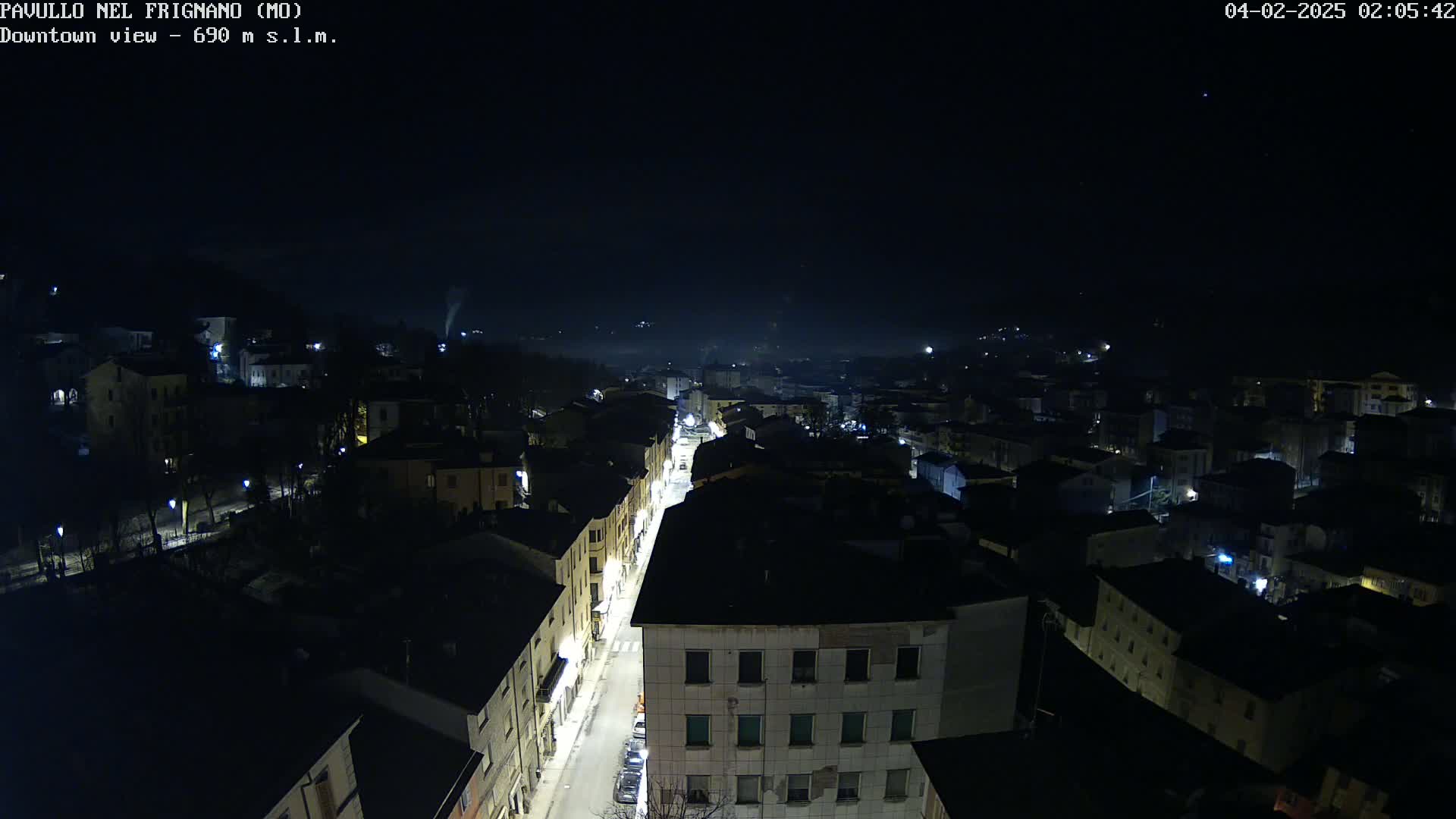 A nighttime aerial view of a town shows a long, illuminated street cutting through a dark, densely packed collection of buildings under a clear, dark sky.