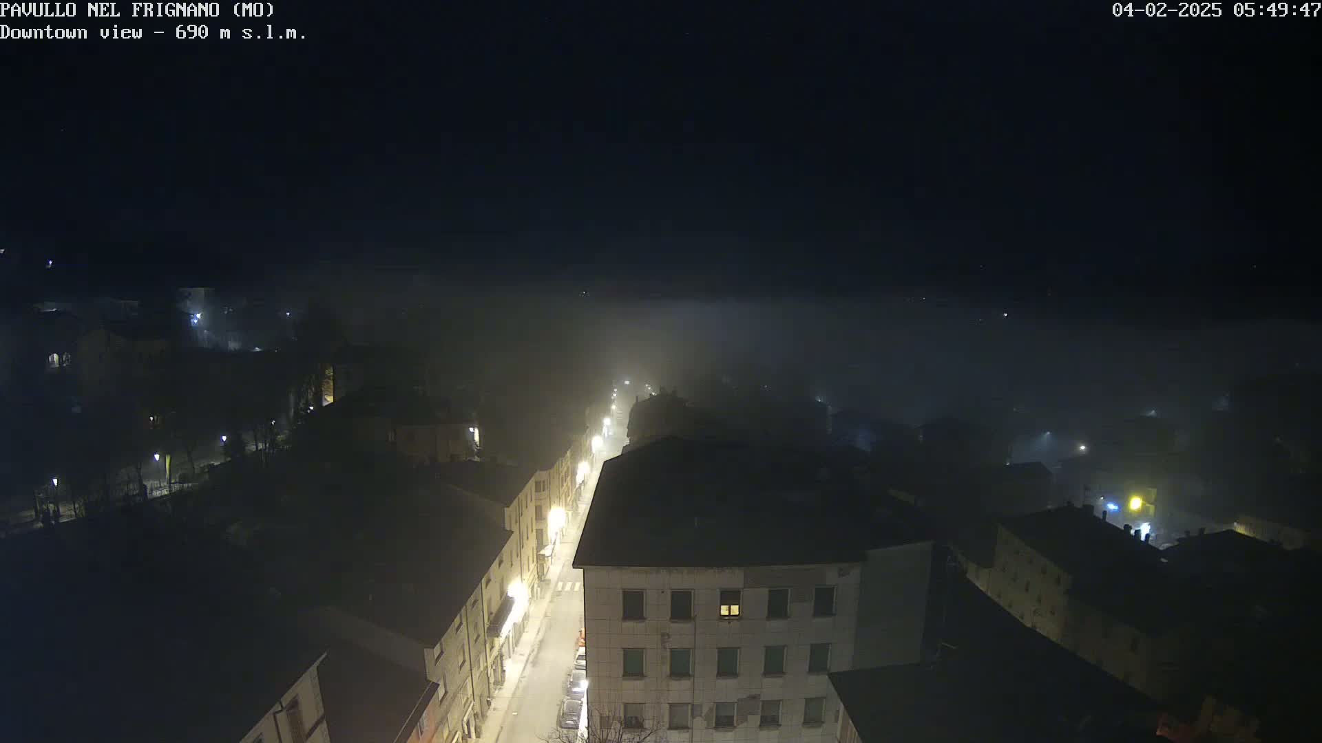 A foggy night scene shows a town's streetlights illuminating buildings and a road below, viewed from an elevated perspective.