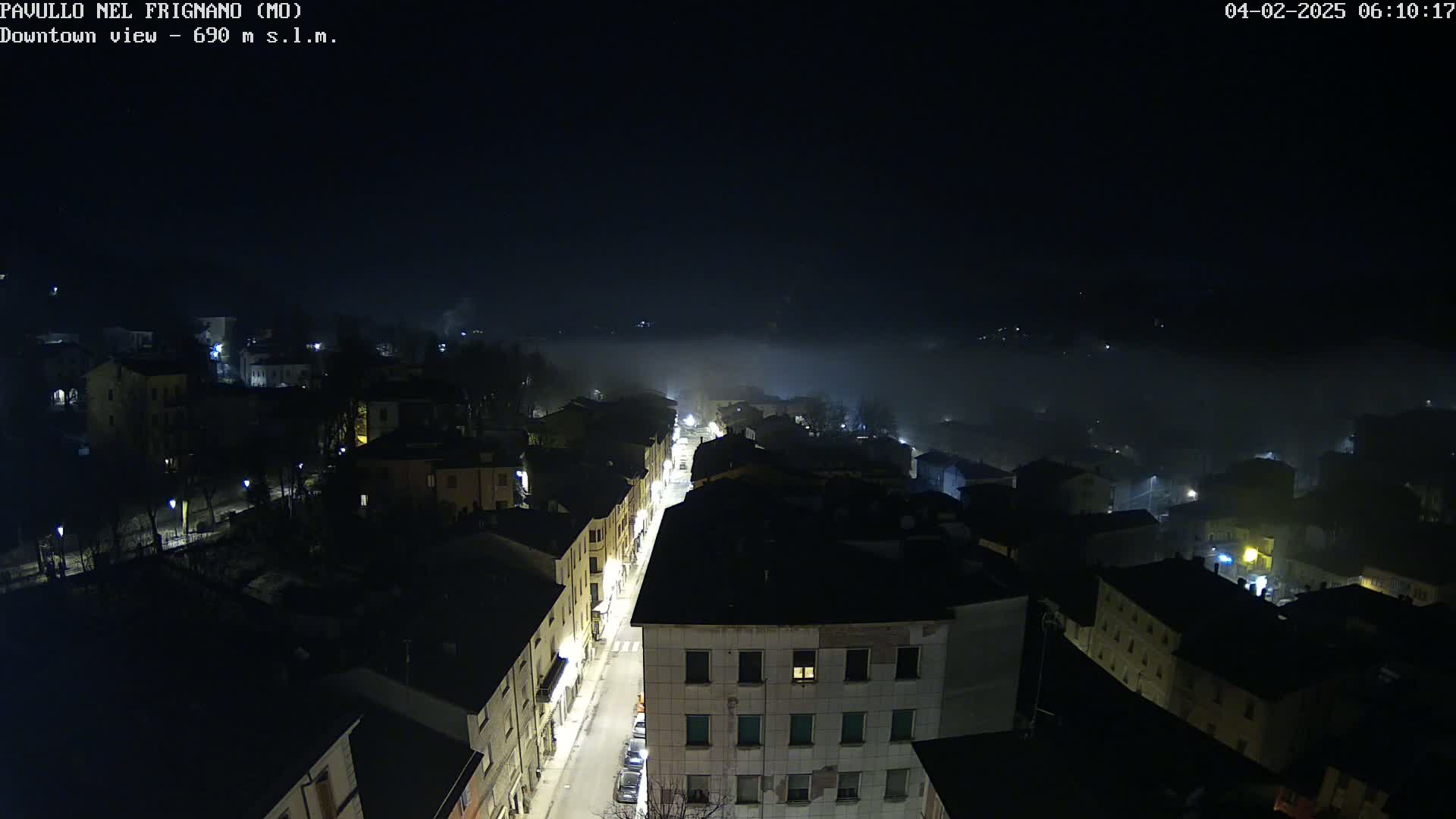 A foggy night scene shows a town's streetlights illuminating buildings and a street below, viewed from an elevated perspective.