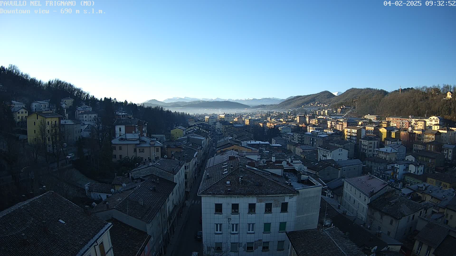 A panoramic view of a town nestled in a valley, under a clear blue sky, with distant mountains partially obscured by fog.