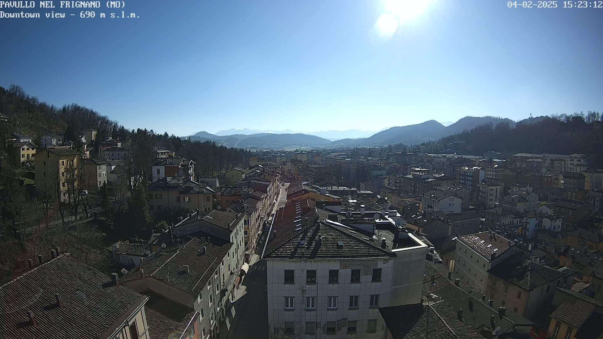 A sunny day reveals a panoramic view of a town nestled in a valley, surrounded by mountains and hills covered in sparse, wintery trees.