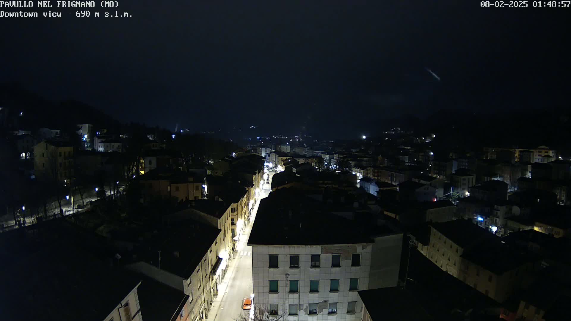 A nighttime, high-angle view of a town with dimly lit buildings and a street with sparse traffic under a dark sky.