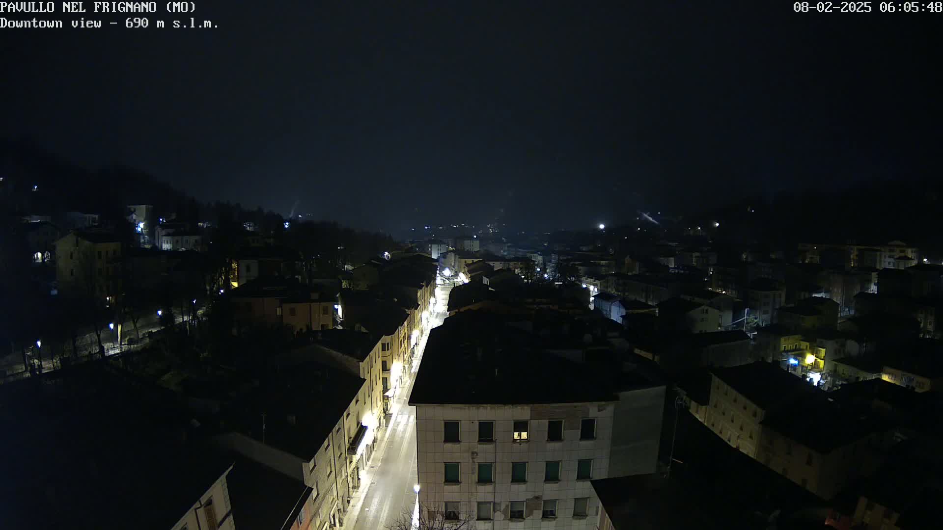 A nighttime, aerial view of a town with dimly lit buildings and a brightly lit street running through its center, under a dark, possibly foggy sky.