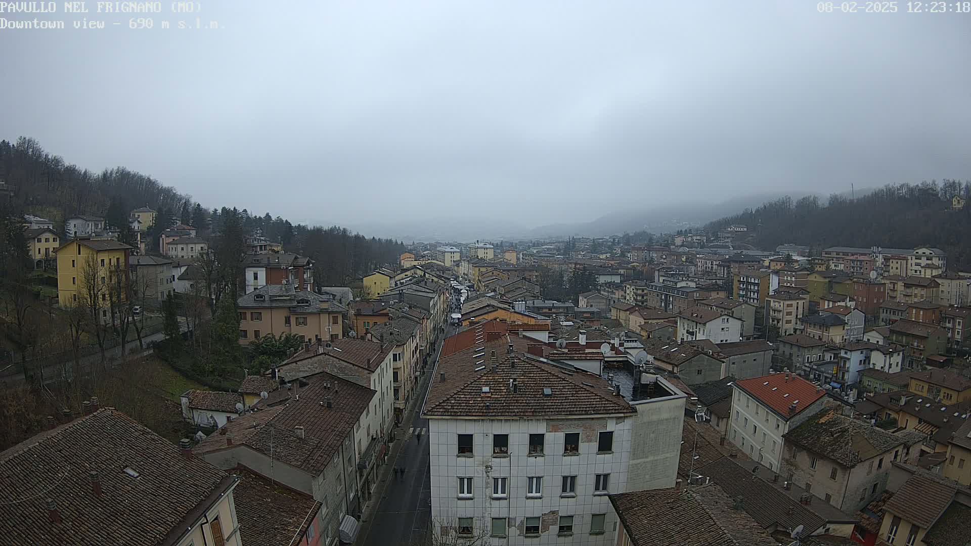 A wide, high-angle view of a town nestled in a valley on an overcast day, showing numerous buildings with red-tiled roofs.