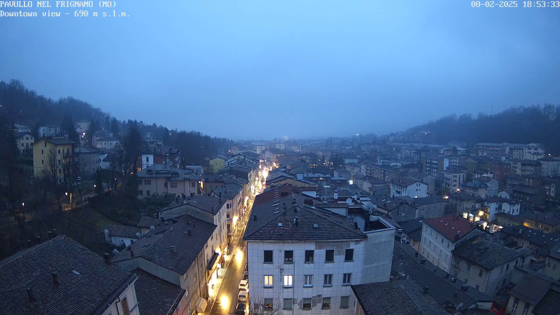 A long street lined with illuminated buildings stretches through a town nestled in a valley under a twilight sky.