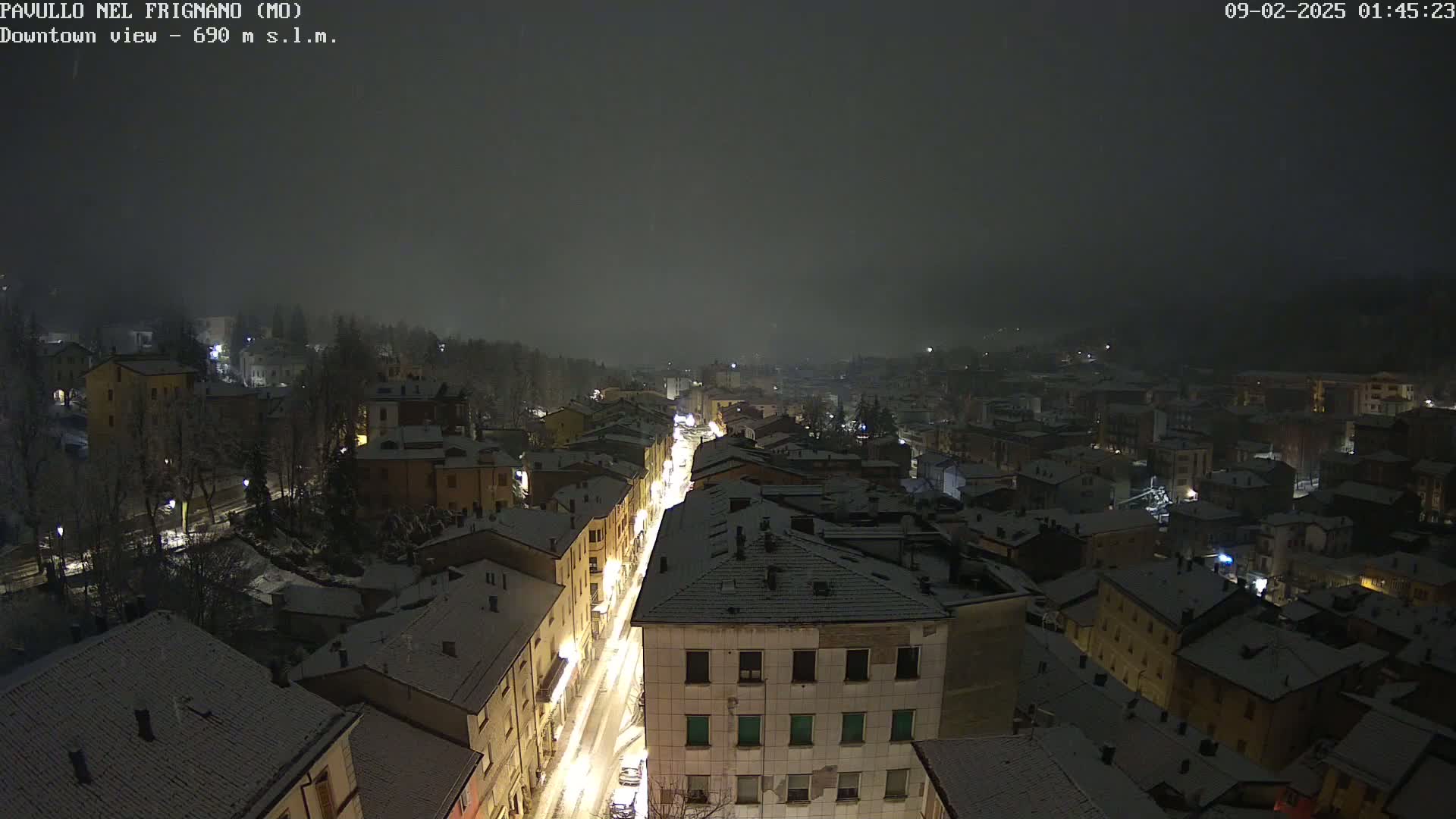 A snow-covered town is viewed at night from above, with lightly falling snow and streetlights illuminating a central street.