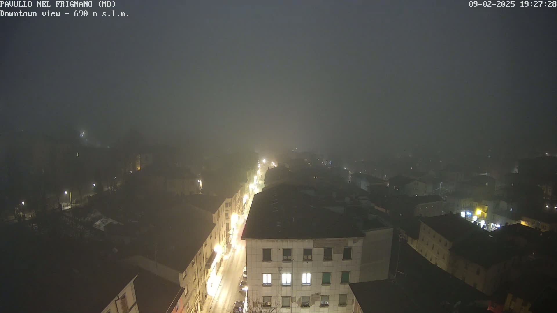 A foggy night scene shows a town's streetlights illuminating buildings and a street below from a high vantage point.
