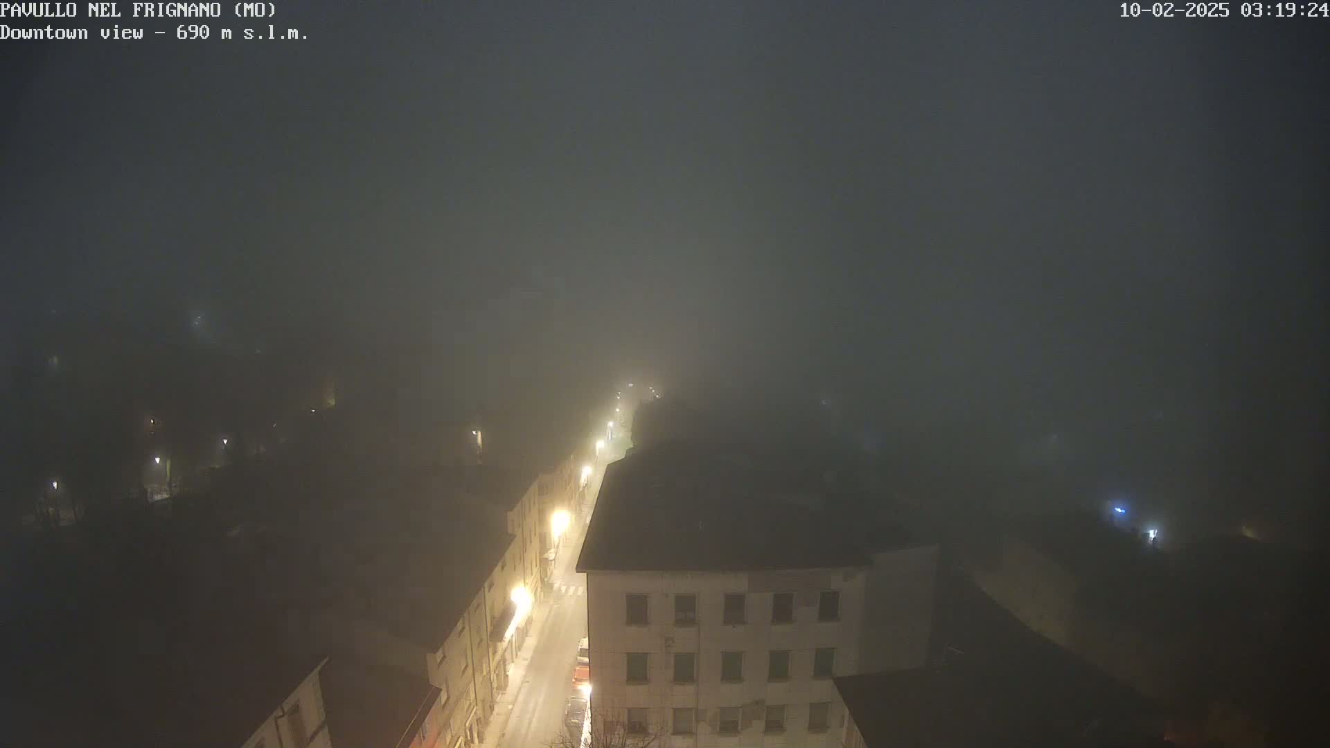 A foggy night scene shows a dimly lit street in a town, with buildings visible through the dense fog.