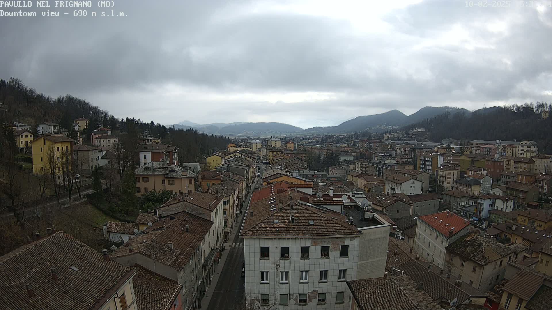 A wide shot of a town nestled in a valley under a cloudy sky, showing many buildings with reddish-brown roofs.