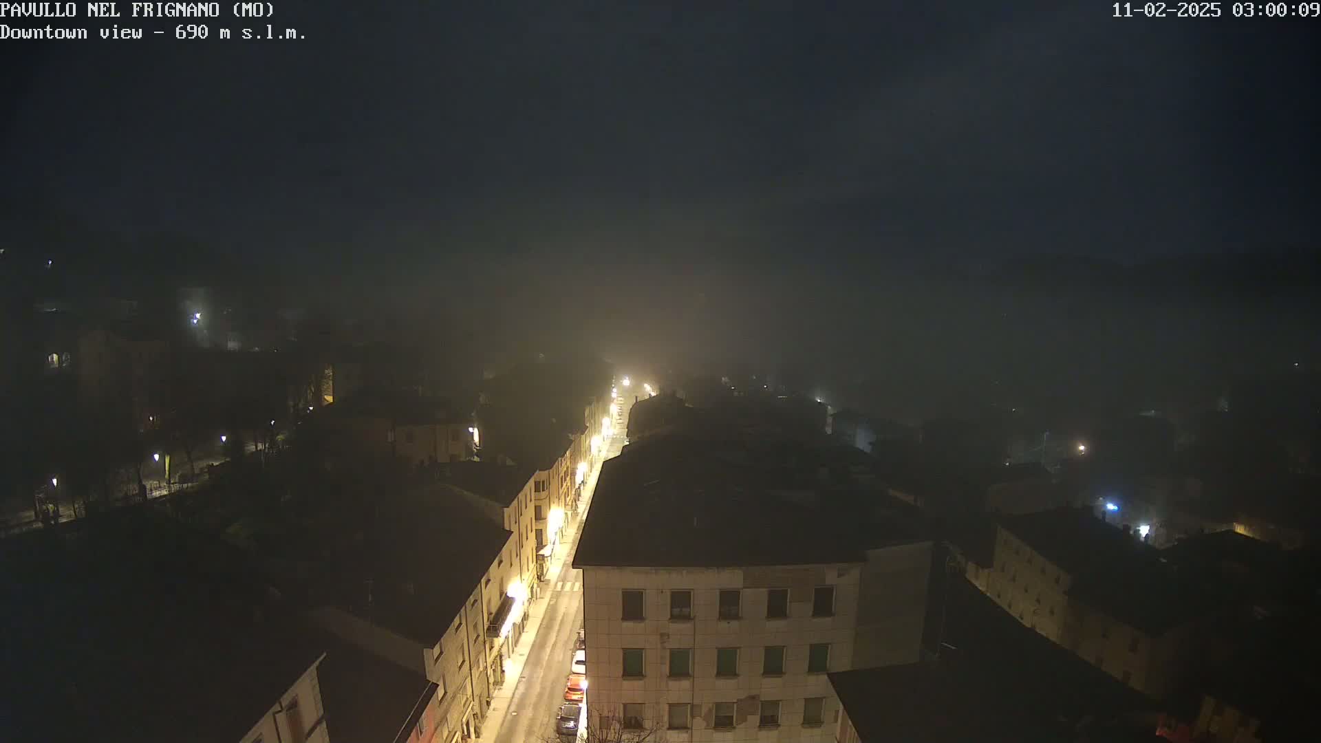 A foggy night scene shows a town's street lit by streetlights, with buildings visible on either side.