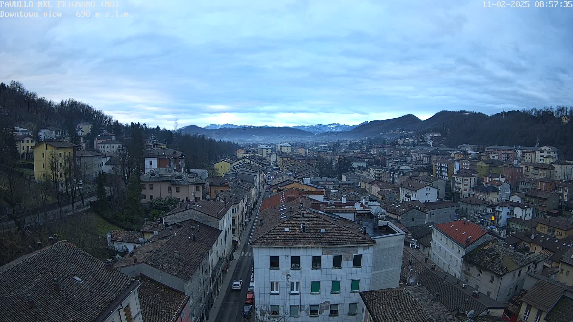 An aerial view shows a town nestled in a valley between tree-covered hills and snow-capped mountains under a mostly cloudy sky.