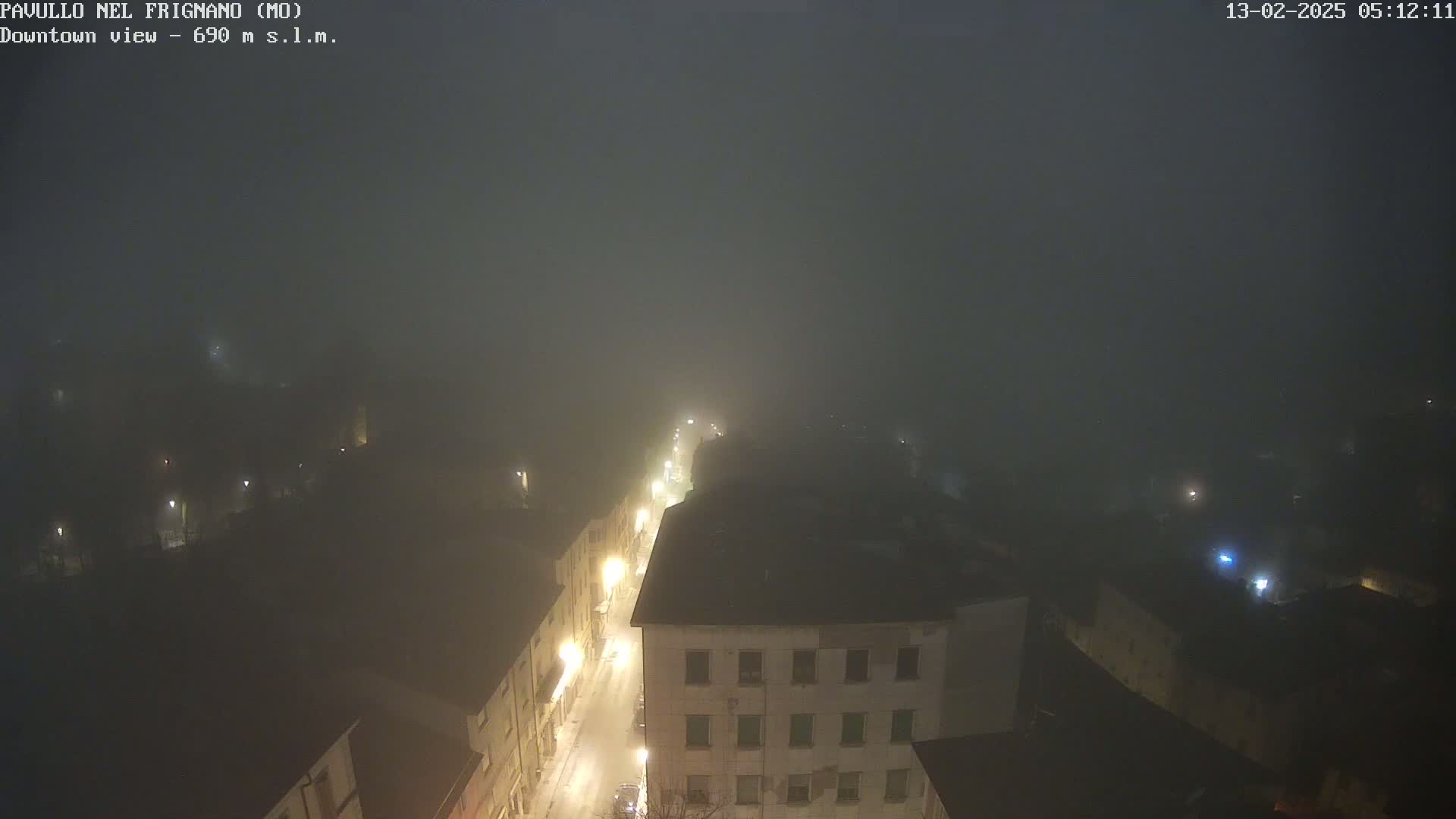 A foggy night scene shows a town's streetlights illuminating a street and buildings below, viewed from an elevated perspective.