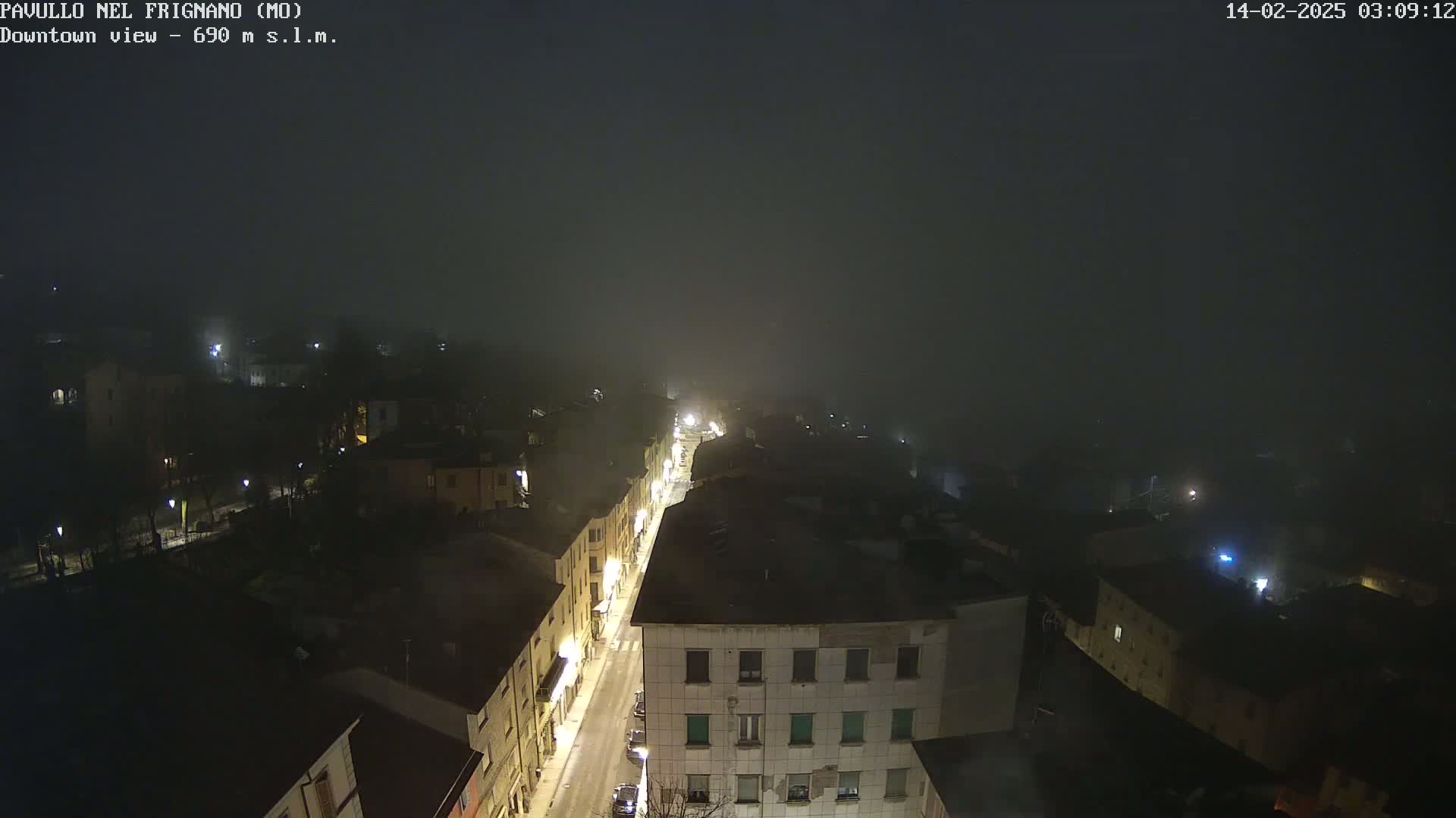 A foggy night scene shows a dimly lit street running through a town, with buildings and streetlights visible through the fog.
