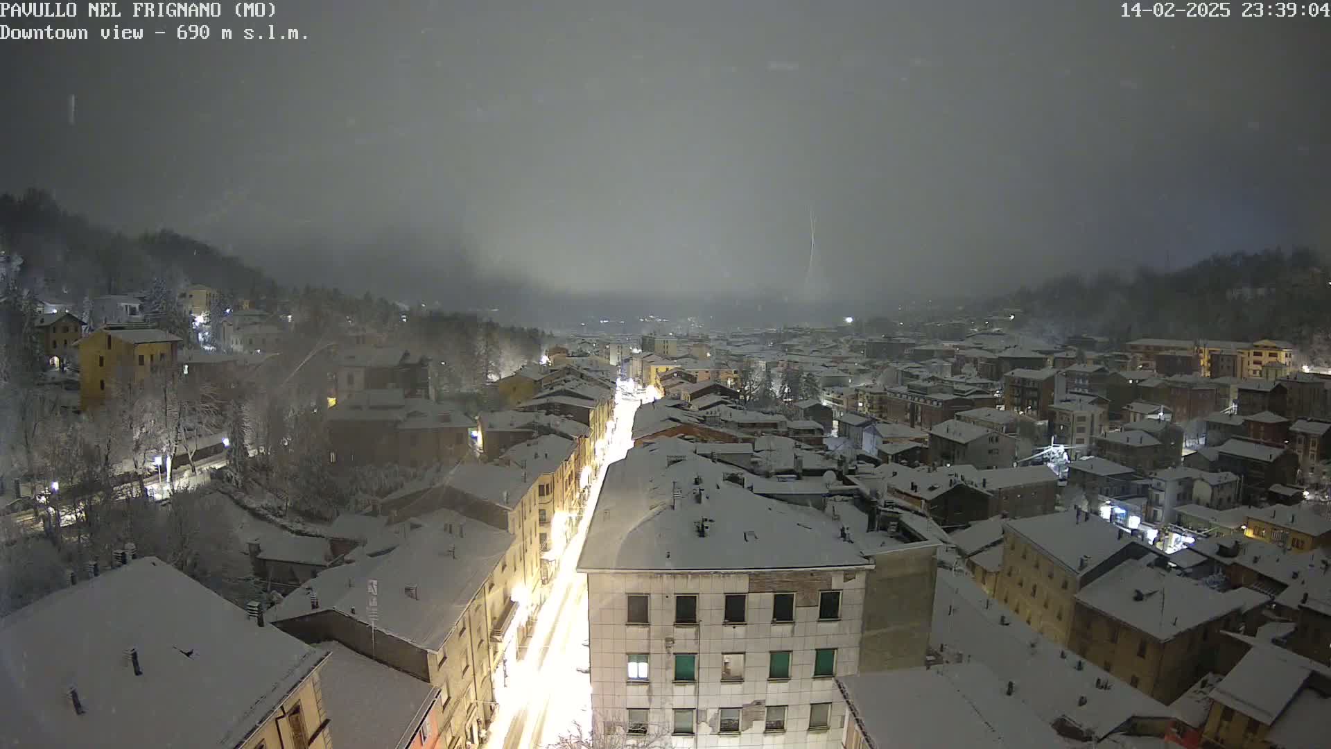 A snow-covered town is viewed at night under a dark, overcast sky, with a brightly lit street visible in the center.