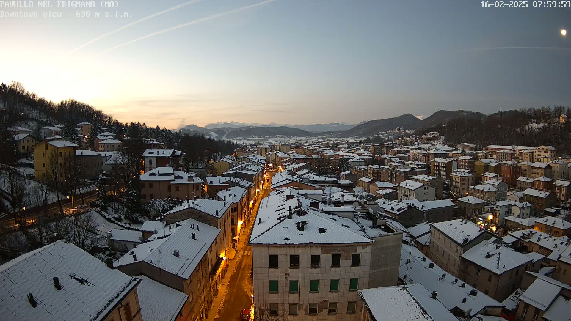 A snow-covered town at twilight, illuminated by streetlights, is nestled in a valley surrounded by dark hills and mountains under a clear sky.