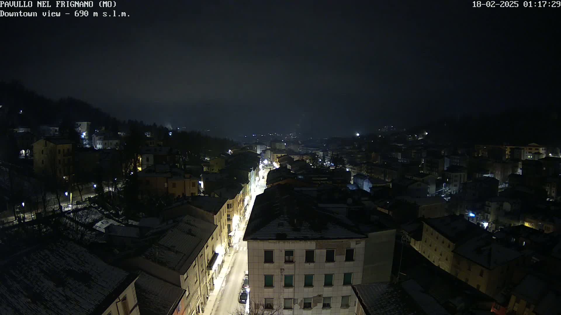 A snow-dusted town at night is viewed from above, with dimly lit streets and buildings under a dark, hazy sky.