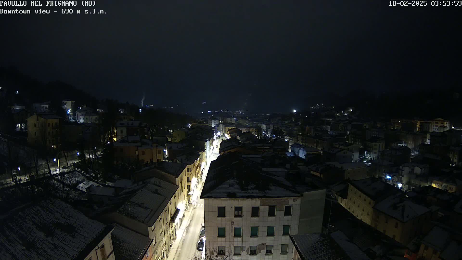 A snow-dusted town at night is viewed from above, showing a long street lined with buildings and illuminated by streetlights under a dark, cloudy sky.