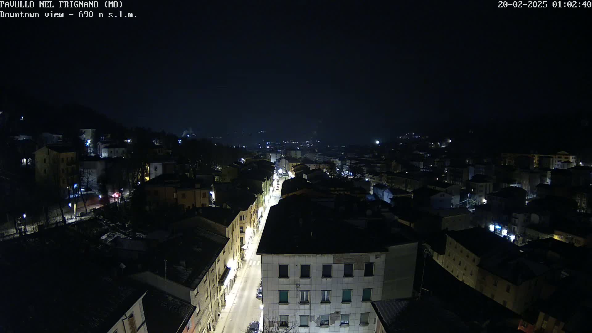 A nighttime, high-angle view of a town with a brightly lit street running through it, surrounded by dark buildings and hills under a clear, dark sky.