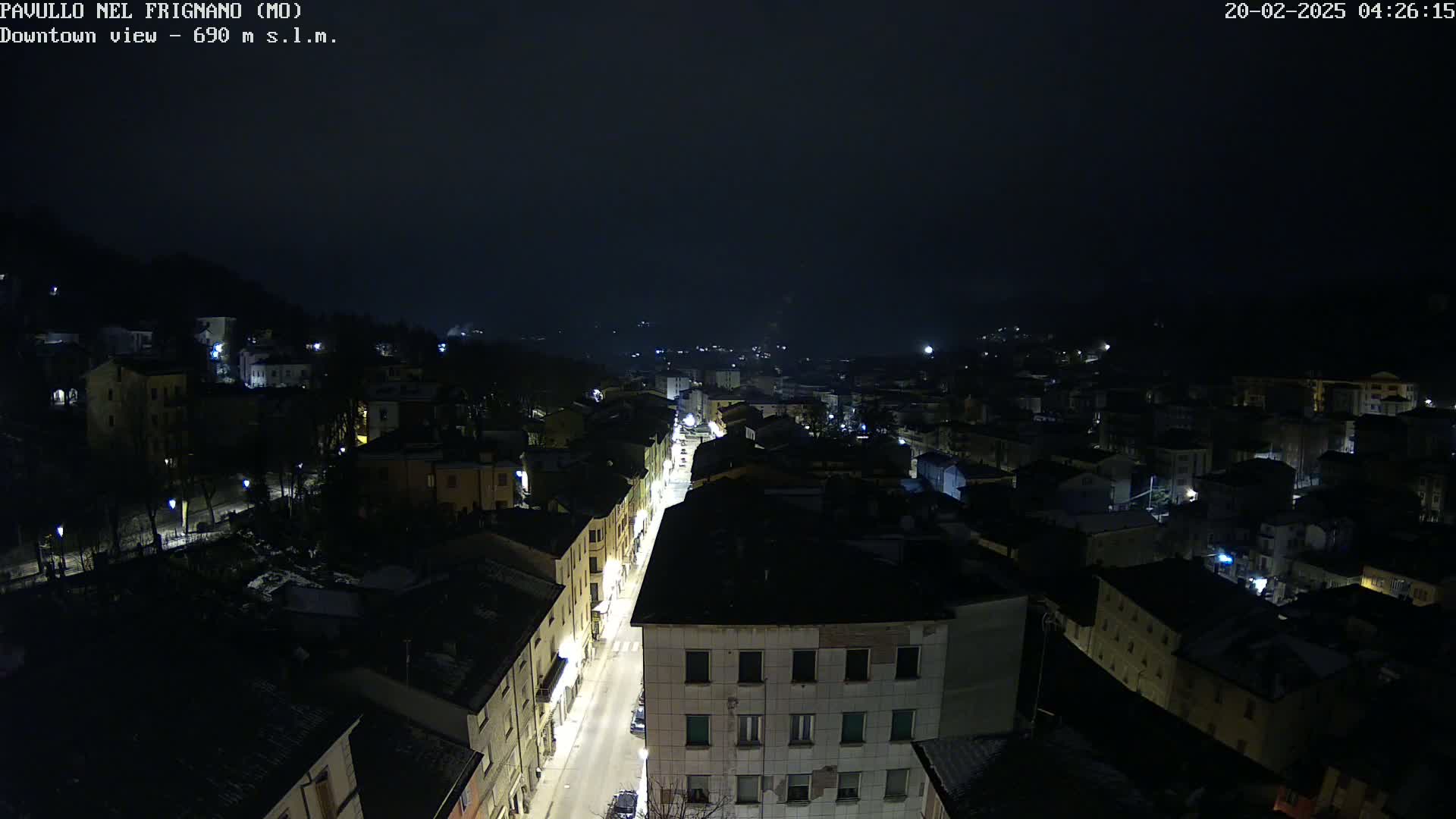 A nighttime, aerial view of a town shows a main street lined with illuminated buildings, surrounded by other buildings in darkness under a dark, overcast sky.