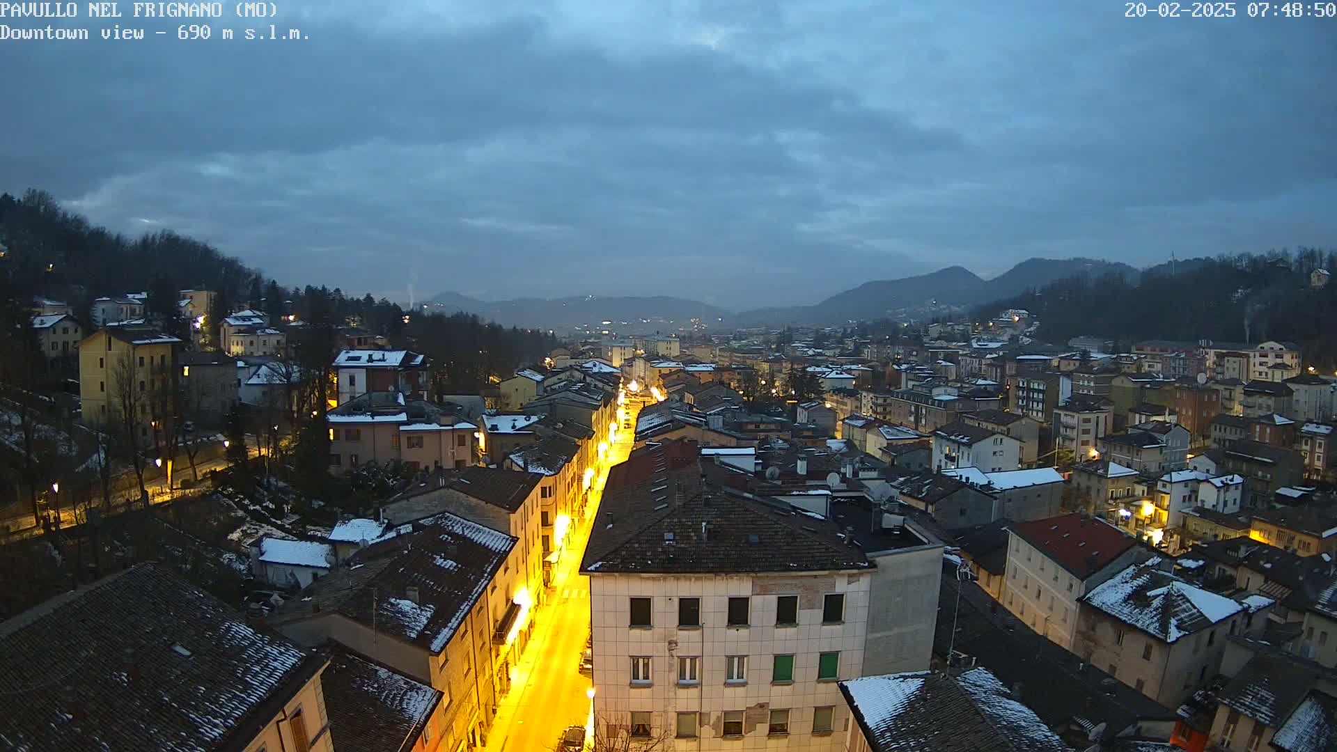 A brightly lit street cuts through a snow-dusted town nestled in a valley under a twilight sky.
