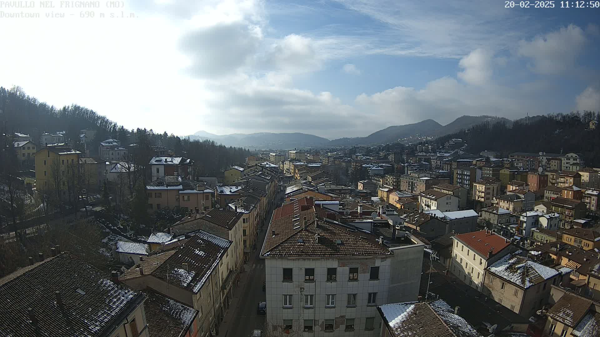 A partly sunny day reveals a sprawling town nestled in a valley between hills, with patches of snow dusting the rooftops.