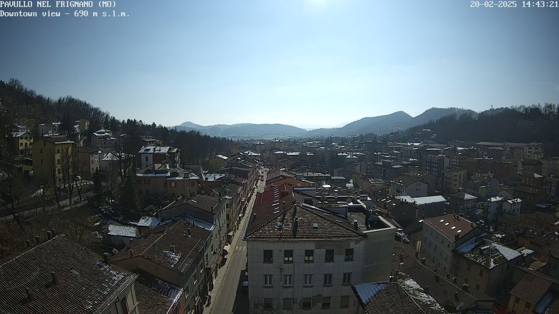 A high-angle, long shot on a sunny day shows a town nestled in a valley between hills, with some snow visible on rooftops and surrounding hillsides.