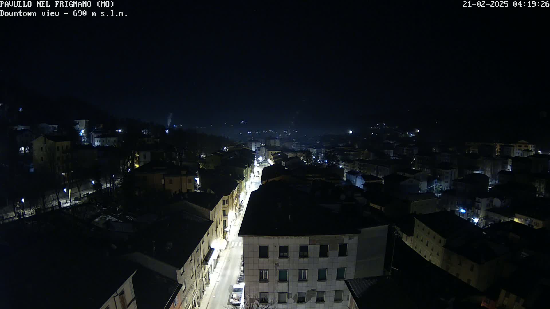 A nighttime aerial view of a town, with dimly lit buildings and a brightly lit street running through the center, under a clear, dark sky.