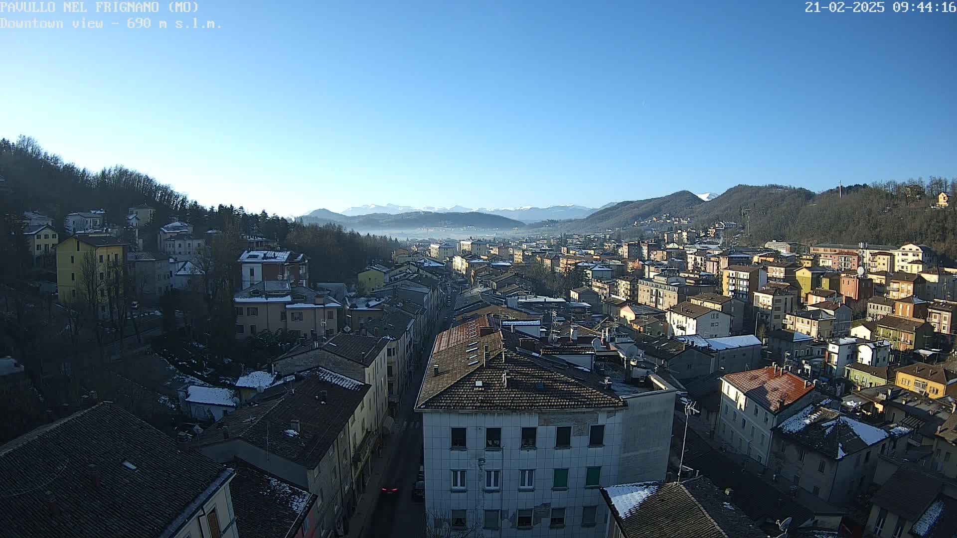 A clear, sunny day reveals a panoramic view of a town nestled in a valley, surrounded by snow-dusted hills and distant mountains.