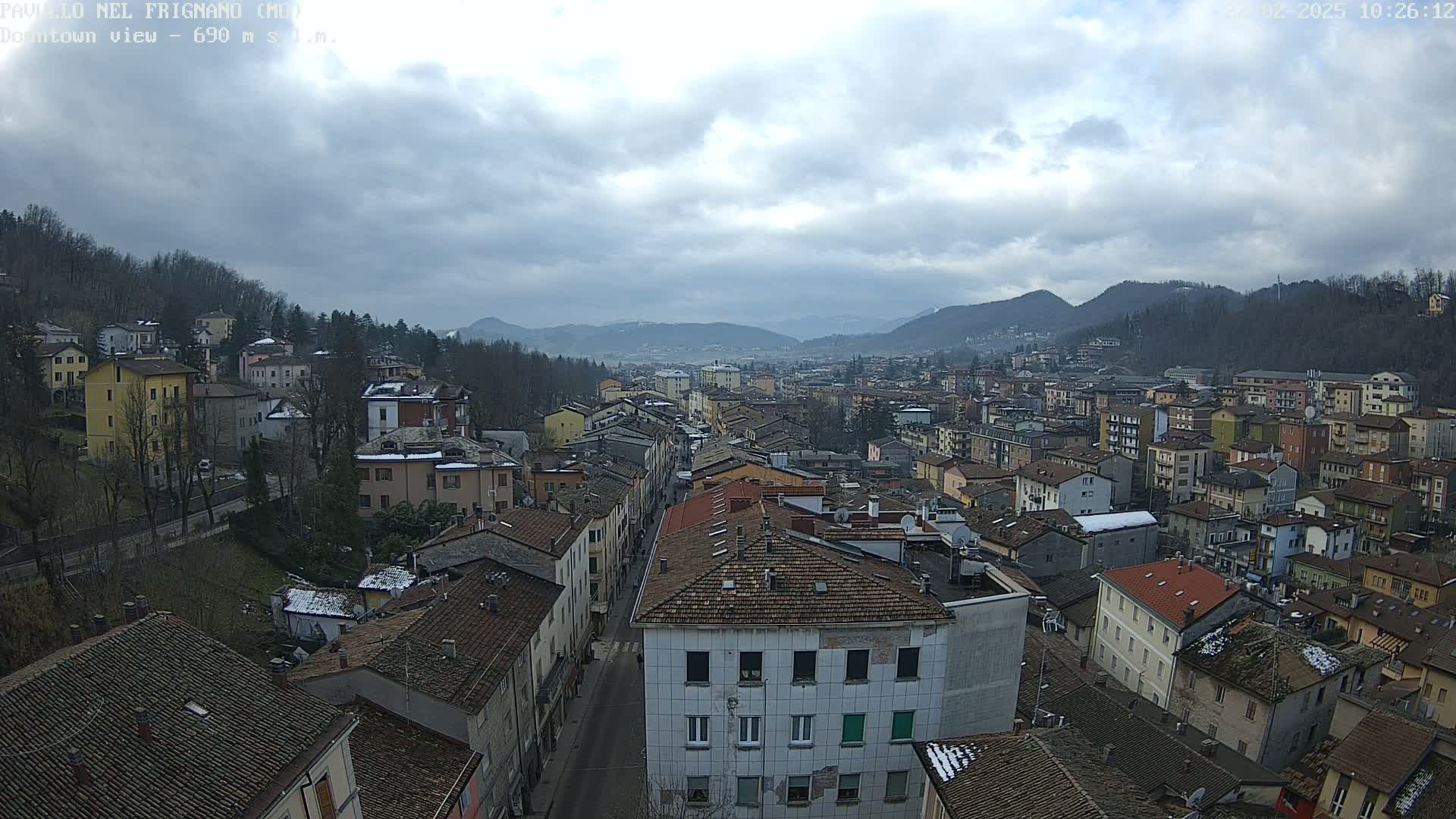 A wide shot of a town nestled in a valley under a cloudy sky, showing numerous buildings with red-tiled roofs and a street running through the center.