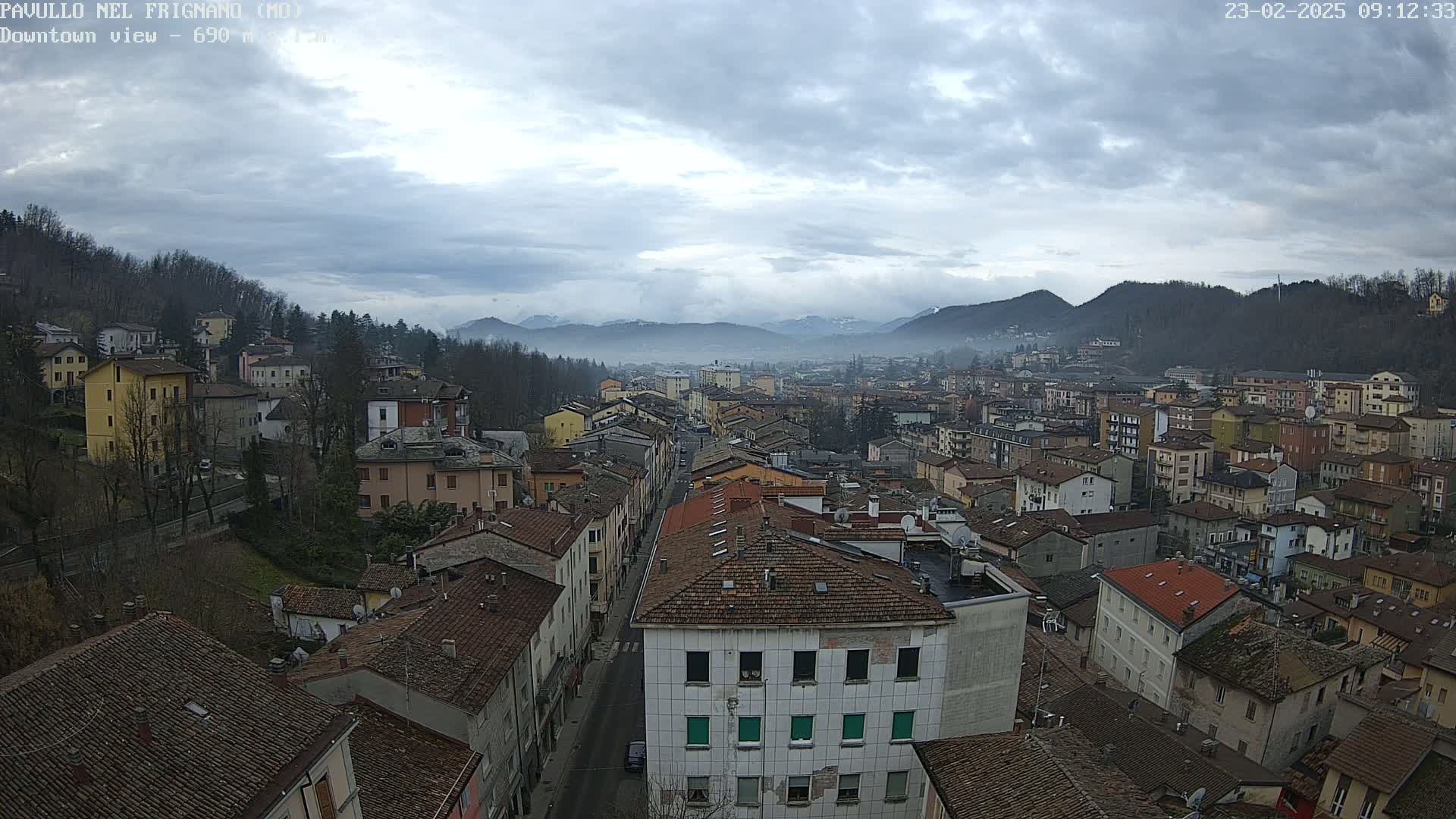 A wide shot of a town nestled in a valley under a cloudy sky, with distant, fog-covered mountains visible in the background.
